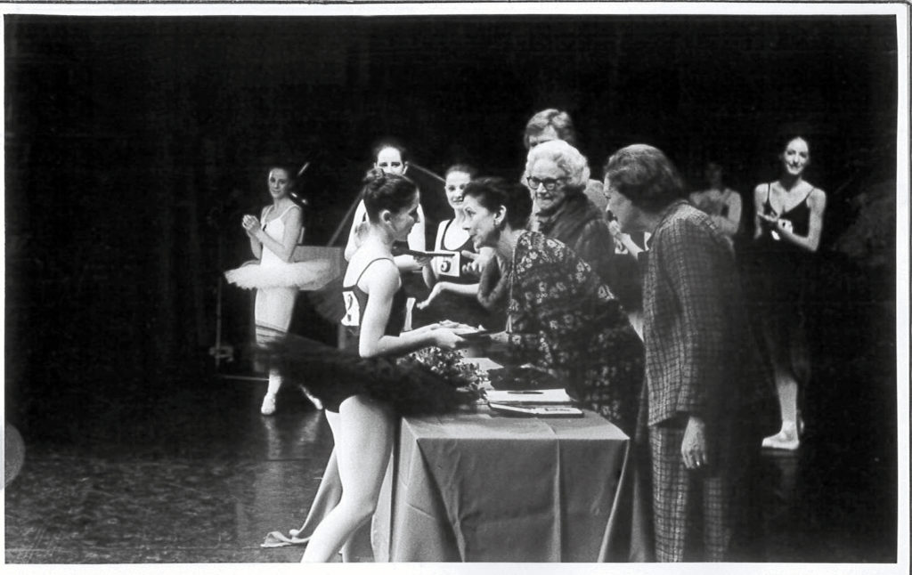 A black-and-white photo of 16-year-old Leanne Benjamin in a leotard and tutu standing on stage accepting her medal from Margot Fonteyn. There is a table in between them and she stands in prep position as she shakes Ms. Fonteyn's hand. Behind them are the rest of the dancers and judging panel. 