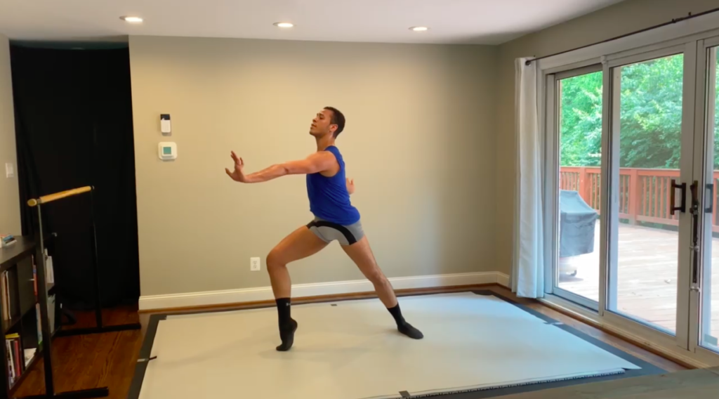 Lucas Castro rehearses on a marley floor taped to his floor at his home. He stands in a large lunge in profile and pushes over his right foot in demi-pointe while stretching his left arm forward and right arm back.