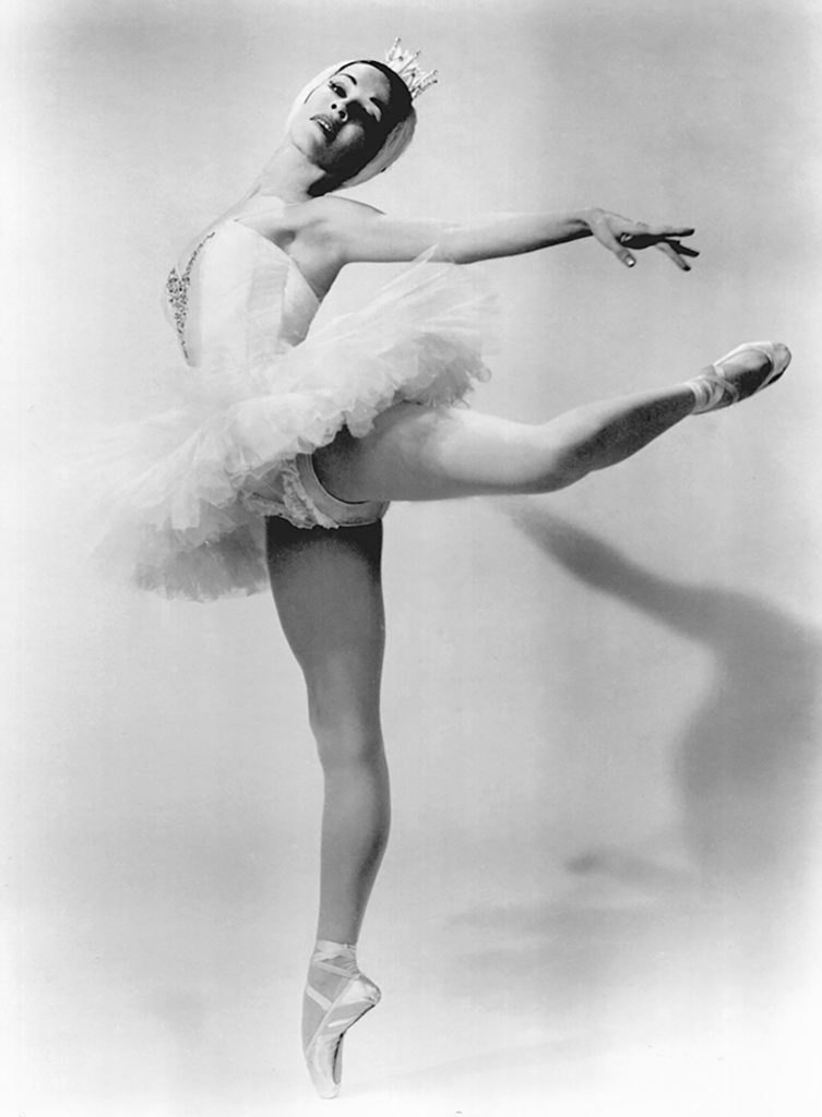 In this black and white photo, Marjorie Tallchief does an attitude derriere in effacé with her left leg up. She arches back with her upper body and looks toward the camera, reaching her arms out and behind her. Tallchief wears a white tutu,tights and pointe shoes and a feathered headpiece with a small tiara.