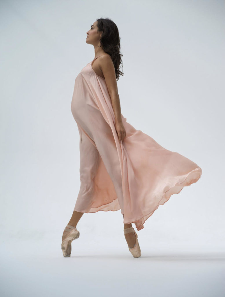 Mathilde Froustey stands in fourth position en pointe in an ankle length pink dress. The dress is blowing back across her body to show her pregnant belly. Her arm are by her side, her dark hair is at her back, and she looks straight forward into the distance.