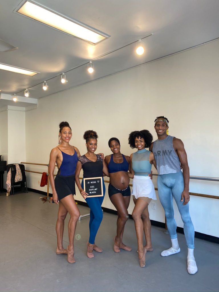 Ingrid Silva stands in a dance studio with two Dance Theatre of Harlem dancers on either side in black bike shorts and a blue sports bra top. The dancer to Ingrid's left holds a snap-on letter sign that says "25 weeks." 
