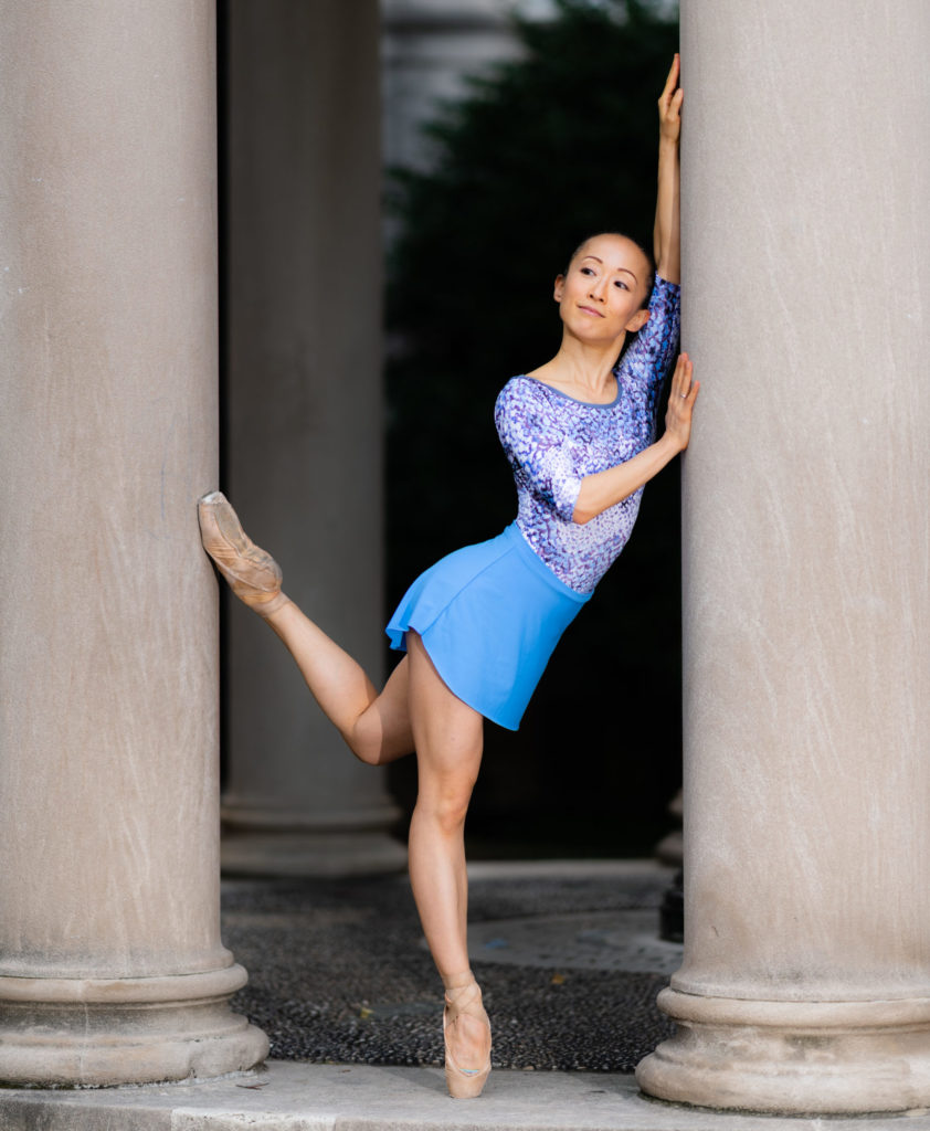 Hope Ruth poses on pointe between two large cement columns. She stands on her right foot, with her left leg bent behind her and rests her head and arms against a column. She wears a blue ballet skirt and blue patterned leotard.