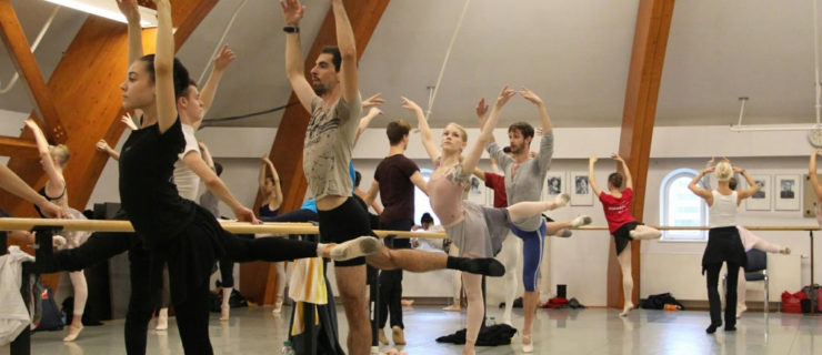 In a large dance studio, a group of male and female summer intensive students in various dancewear balance on their left foot in an attitude croisé at the barre.