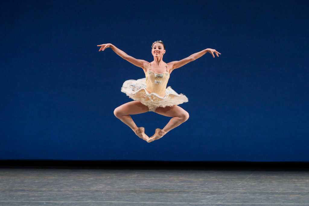 Costumed in a light yellow tutu, Tiler Peck performs a large pas de chat onstage with her arms out in second position. She dances in front of a blue backdrop.