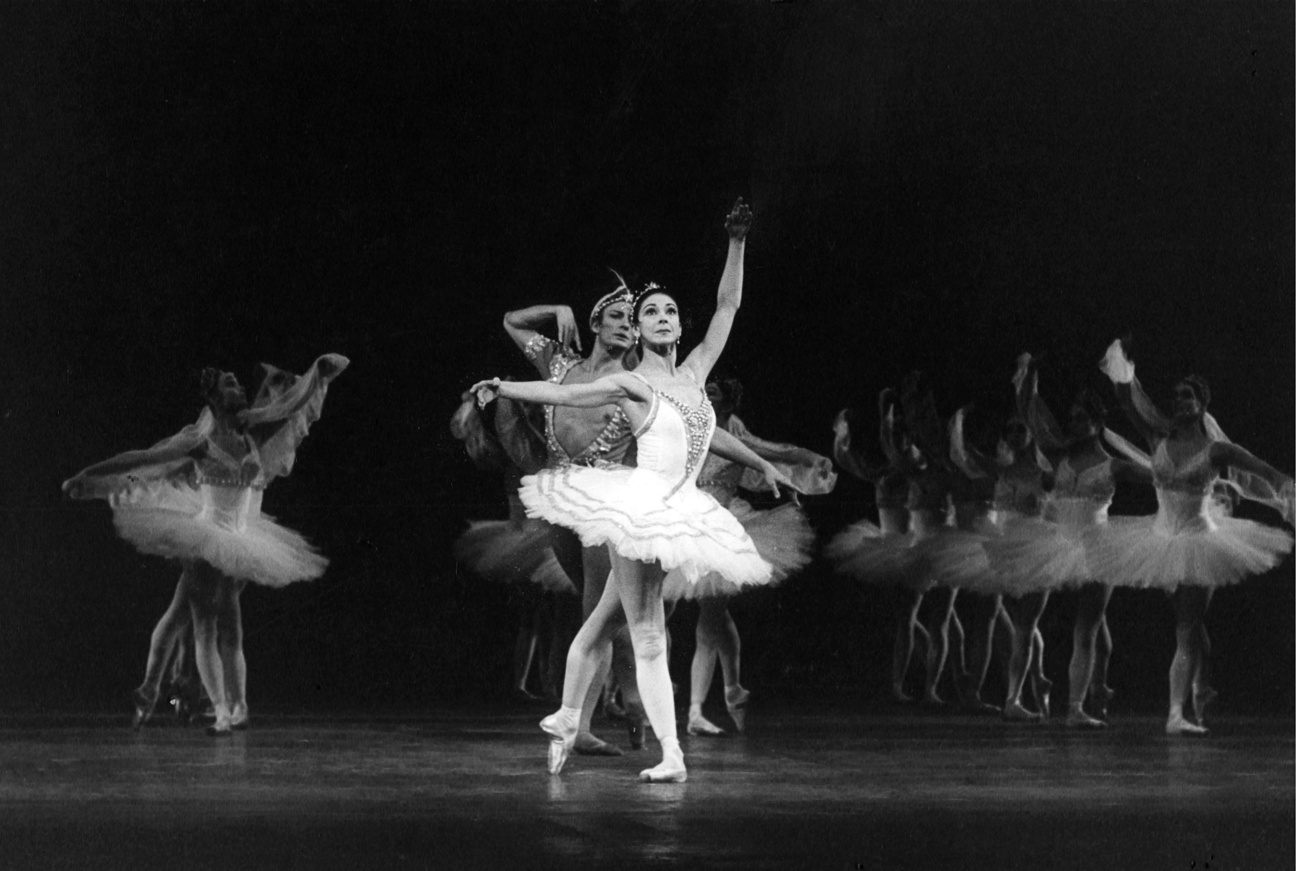 A black and white photo of Margot Fonteyn standing at the front of the Royal Opera House stage. She wears a white tutu and looks out into the audience with her arms in fourth position and her back leg pointed. Behind her is Keith Rosson, her partner for "La Bayadere," and three columns of corps de ballet dancers all in the same position as her.