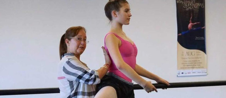 Magaly Suárez, wearing a long-sleeved blouse and leggings, presses her right hand against Suttyn SImon's back to correct it while she balances in retiré. Suttyn wears a pink leotard, pink tights and pointe shoes, and a black skirt.