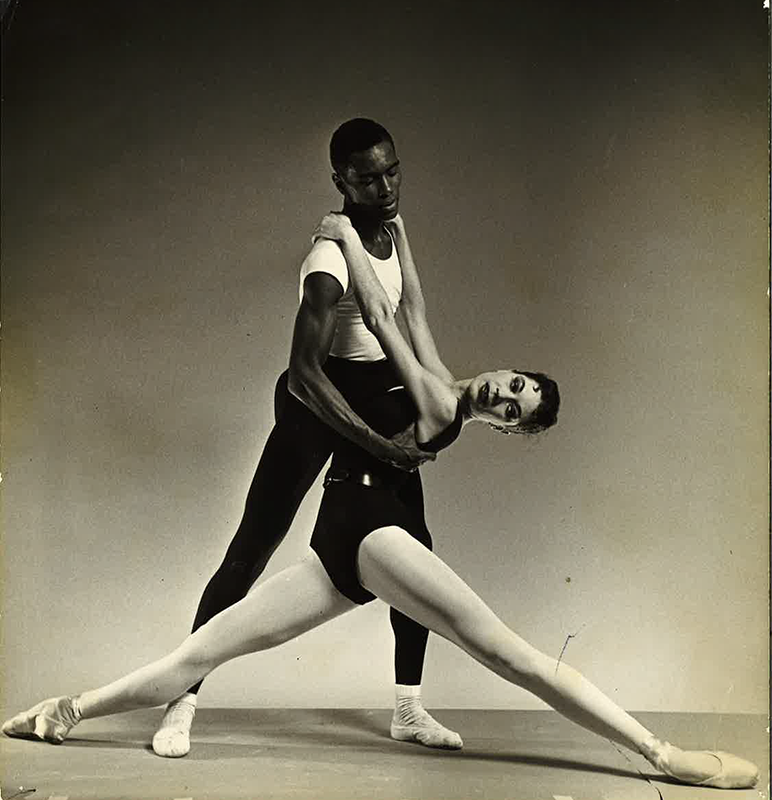 In this black and white photo, Arthur Mitchell holds onto the shoulders of Diana Adams as she lowers into a split, looking over her left shoulder. Adams wears a black leotard, pink tights and pointe shoes, while Mitchell wears a white T-shirt, black tights, white socks and white ballet slippers.