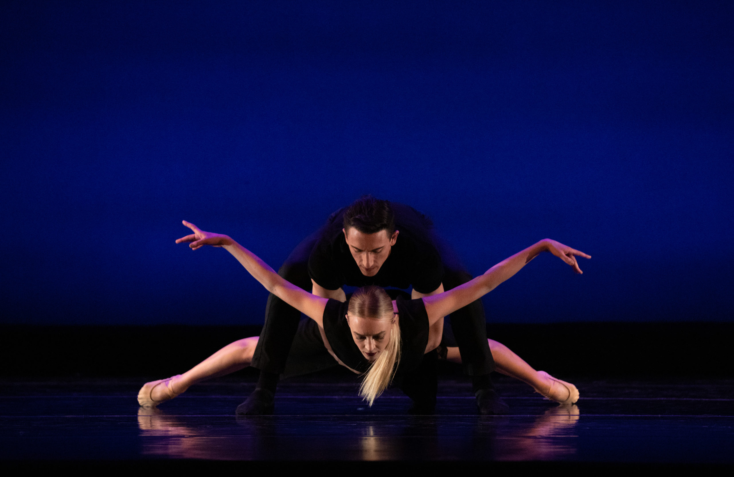 Dance Aspen dancer Anthony Tiedeman crouches forward onstage and holds Kaya Wolsey around the waist. Kaya is close to the floor and spreads her arms and legs out to the side. Anthony wears a black shirt and pants, while Kaya wears a short black dress.