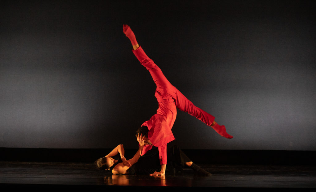 Dance Aspen dancer Katherine Bolaños lays on the stage floor with her knees bent and feet on the ground and presses her right hand into a male dancer's head. The man, in red pants and red shirt, balances on his hands and splits his legs above her.