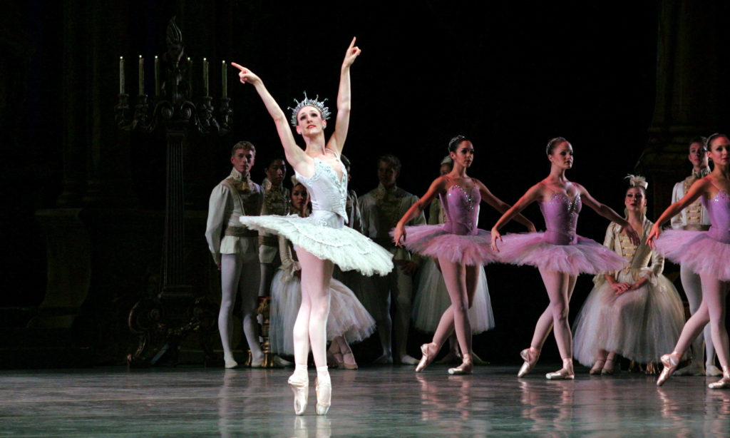 Tricia Albertson wears a white tutu, crown and pointe shoes and stands onstage in sus-sous with her arms lifted high in a V-shape. She points her fingers to the sky and looks up. Behind her onstgae a line of corps dancers in purple tutus stand in B plus position.