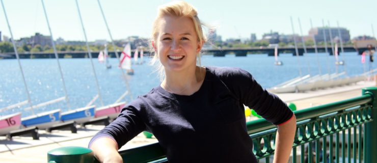Grace Young, wearing a dark, long-sleeved Tshirt, leans against a railing at a marina and smiles cheerfully at the camera. Behind her is a river with several small sailboats.
