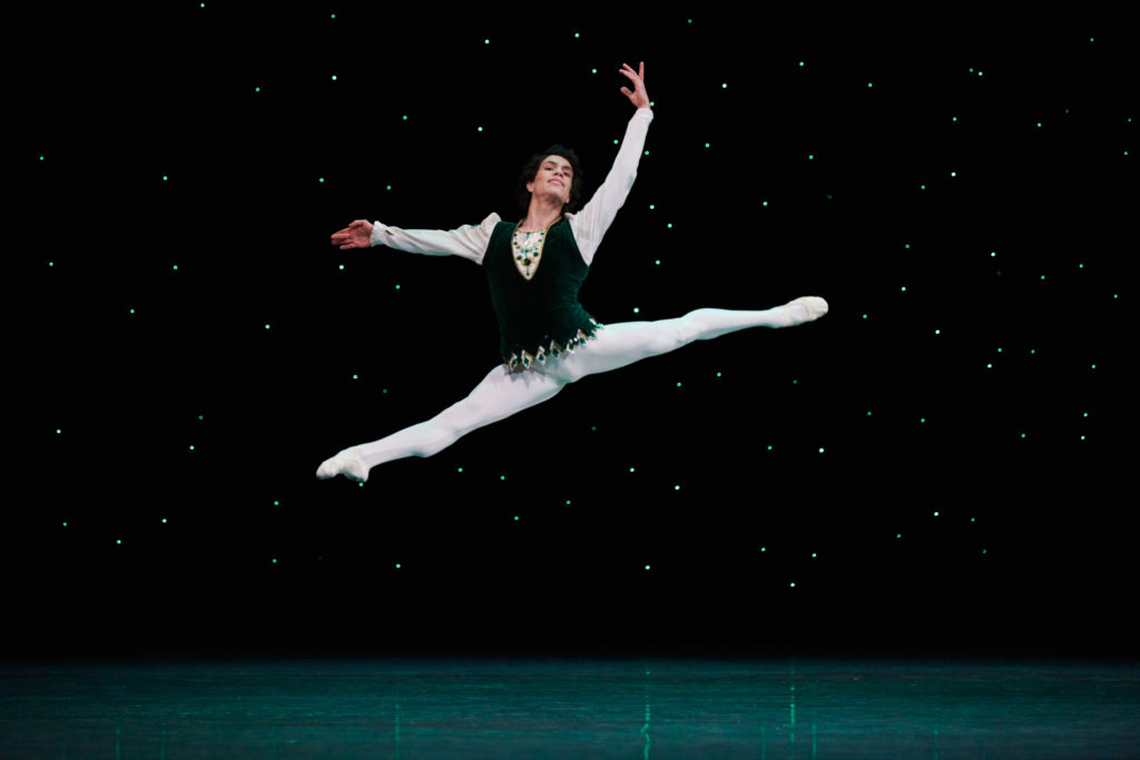 Esteban Hernández performs a large saut de chat onstage, in front of a dark, starry backdrop. He wears a green velvet tunic with white sleeves and white tights and ballet slippers.
