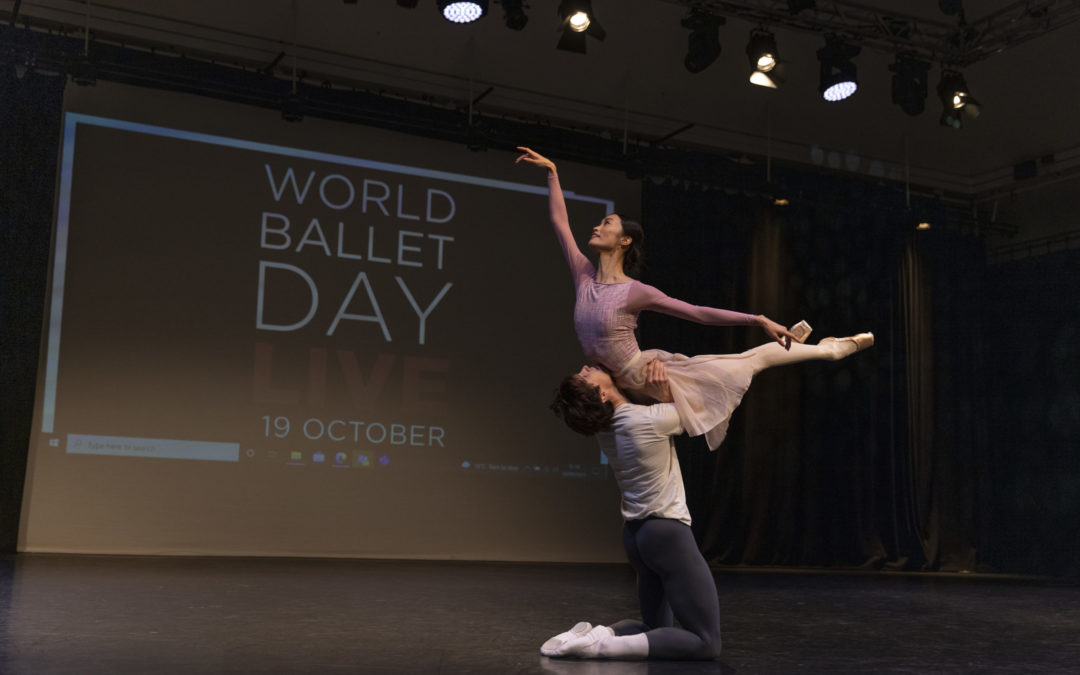 William Bracewell, kneels on the ground and holds Fumi Kaneko overhead by the hips as she arches her upper body up and lifts both her legs behind her. Both wear dance clothing and dance onstage in front of a backdrop that says World Ballet Day LIVE 19 October.
