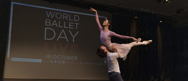 William Bracewell, kneels on the ground and holds Fumi Kaneko overhead by the hips as she arches her upper body up and lifts both her legs behind her. Both wear dance clothing and dance onstage in front of a backdrop that says World Ballet Day LIVE 19 October.