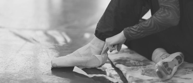 A ballerina sits on the floor in the theater's rehearsal hall and puts on pointe shoes.