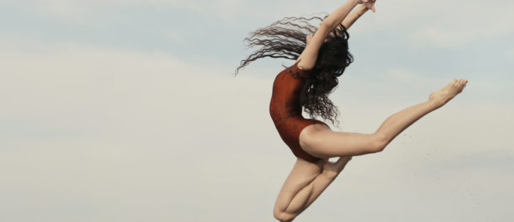 A ballerina in a bronze-colored leotard jumps in the ocean, tucking her right leg up and her kicking back leg in attitude. She arches back with her arms in high fifth and her with long, curly hair flying free.