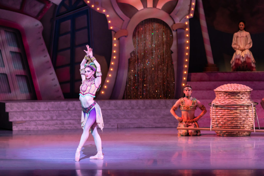 Julia Eisen stands downstage with her front leg popped in front of her and her hands held together overhead. She wears a costume for the Arabian divertissement of the Nutcracker. Behind her a man kneels upstage at the base of an opulent set.  