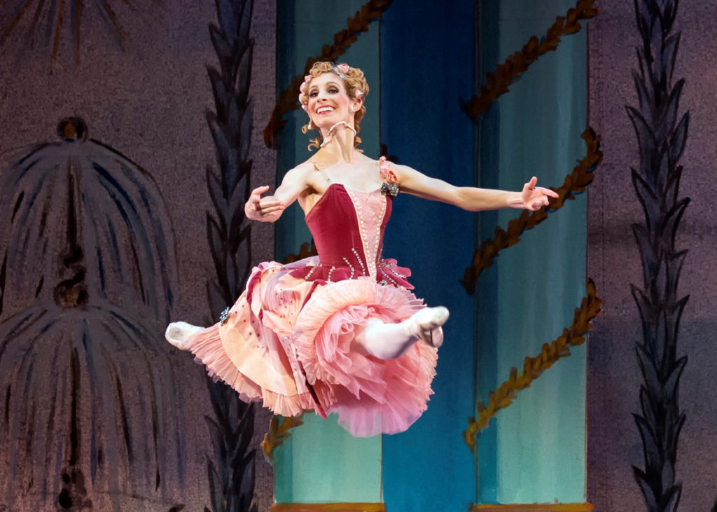 Kate Honea executes a grand jeté en avant in attitude during an onstage performance. She wears a pink and rose colored costume with a long tutu, velvet bodice and floral headpiece.