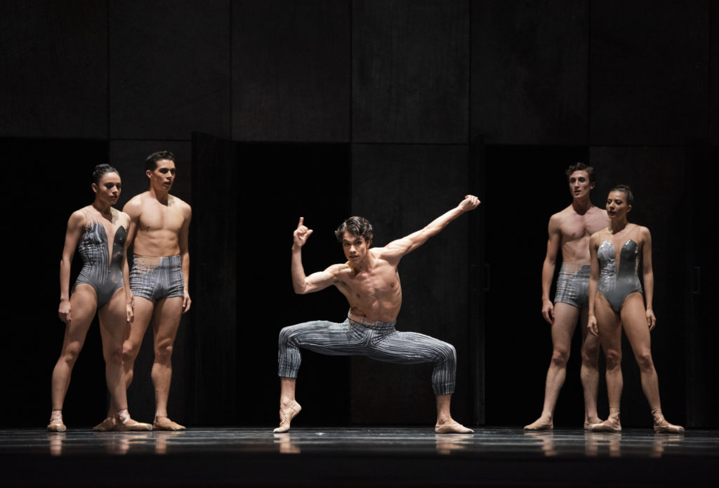 Esteban Hernández, wearing gray and black patterned tights, does a deep second position plié, propping his right foot onto demi-pointe on a darkened stage. Two couples in similar costumes stands behind and to the side of him.