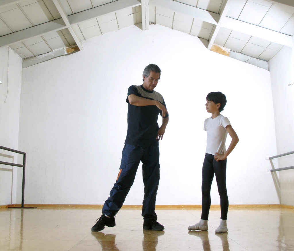 Hector Hernandez, in sneakers and workout clothes, stands to the left of his young son Esteban and shows him a dance move. Esteban, wearing a white T-shirt, black tights and ballet shoes, stands with his hands on his hips and watches intently.