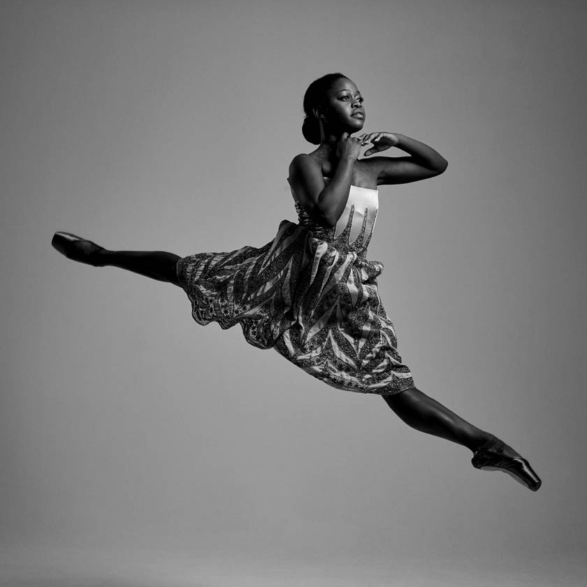 In this black and white photo, ballerina Michaela DePrince performs a sissone fermé and holds her hands just under her chin. She wears a sleeveless dress with a white top and patterned skirt.