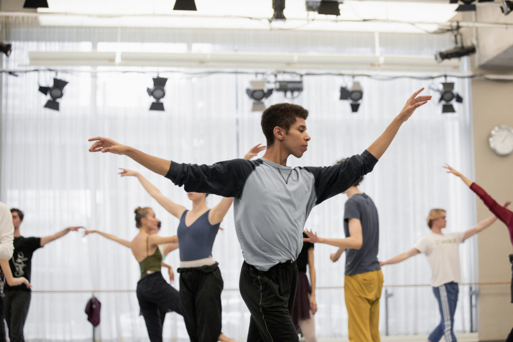 Alexander Skinner stands at the front of a dance studio with his hands lifted to the height of his shoulders. He wears a loose-fitting long sleeved grey/black shirt and dark pants. Behind him are other dancers in rehearsal garb mirroring a similar position. 