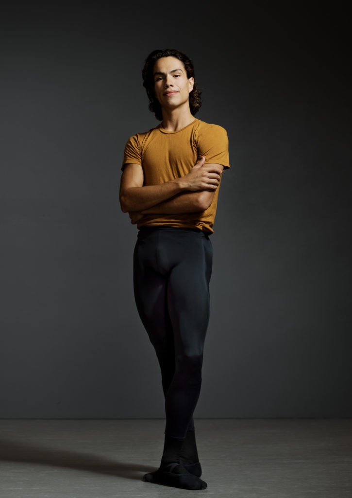 Esteban Hernández stands regally with his arms crossed in front of his chest and with a confident smile. He wears a gold T-shirt and black tights, black ballet shoes.