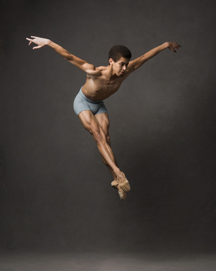 Alexander Skinner is suspended mid-jump against a dark monochrome background. His legs are held together and his body bends forward at the waist, with his arms out behind him. He wears no shirt and short grey shorts.