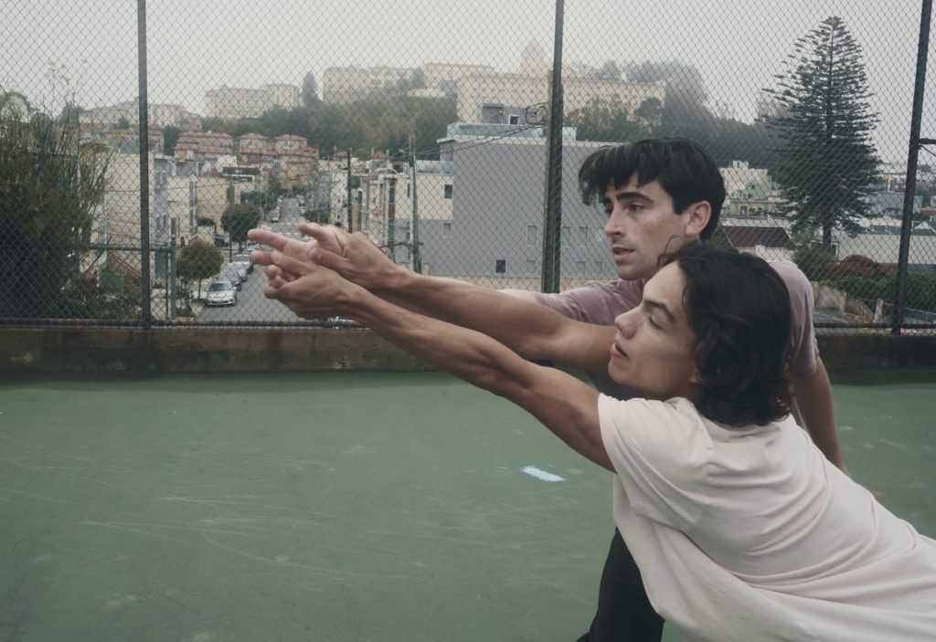 Esteban Hernández and Joseph Walsh lunge forward together with their arms outstretched in a parallel direction. They stand on an outdoor sports court that is made of green concrete and are framed by the foggy San Francisco skyline. Hernández wears a white t-shirt, Walsh wears red. 