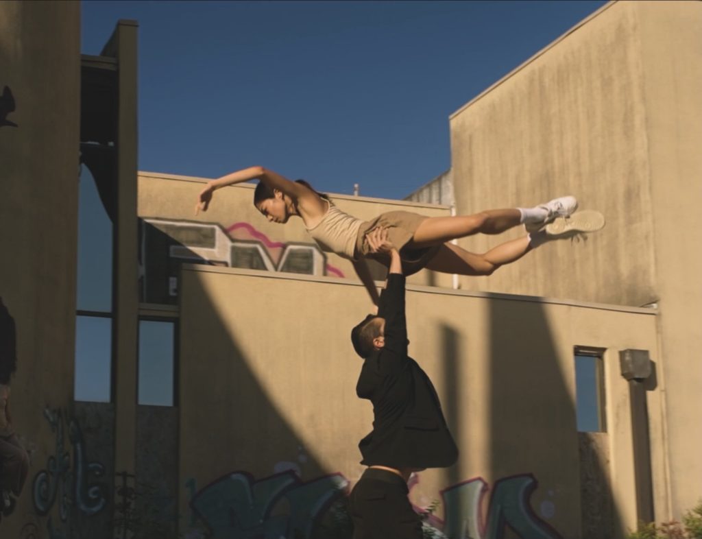 A male dancer holds a female dancer horizontally over his head. They stand outside surrounded by concrete buildings and alternating stripes of sunlight and shadow. He wears a dark shirt and pants, she wears tan shorts and a tank top, with white sneakers on her feet. 