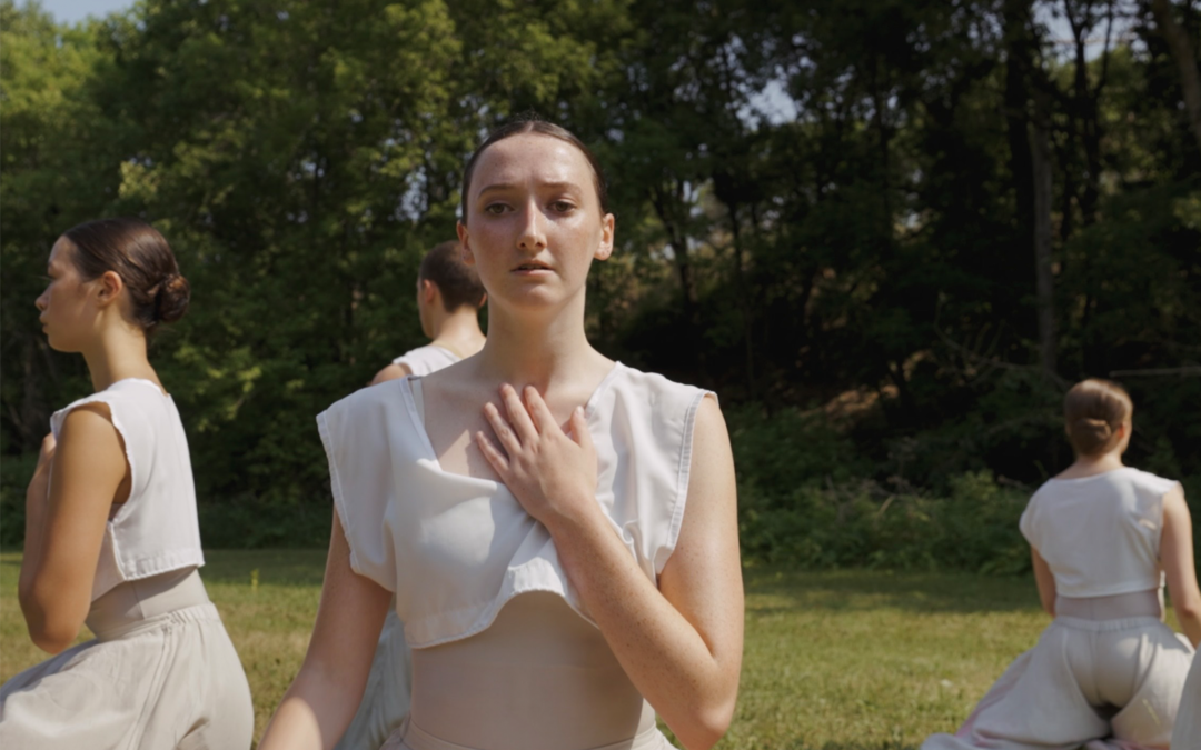 Maggie Weatherdon kneels on the ground with her left hand over her chest. She sits in the middle of a grassy meadow and wears a flowing dress made of white and mauve fabric. Behind her, several other female dancers mirror the position in different directions.