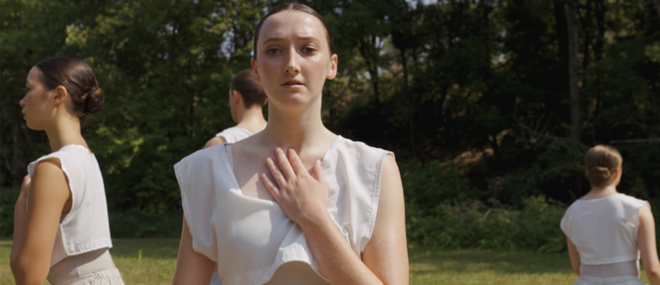 Maggie Weatherdon kneels on the ground with her left hand over her chest. She sits in the middle of a grassy meadow and wears a flowing dress made of white and mauve fabric. Behind her, several other female dancers mirror the position in different directions.