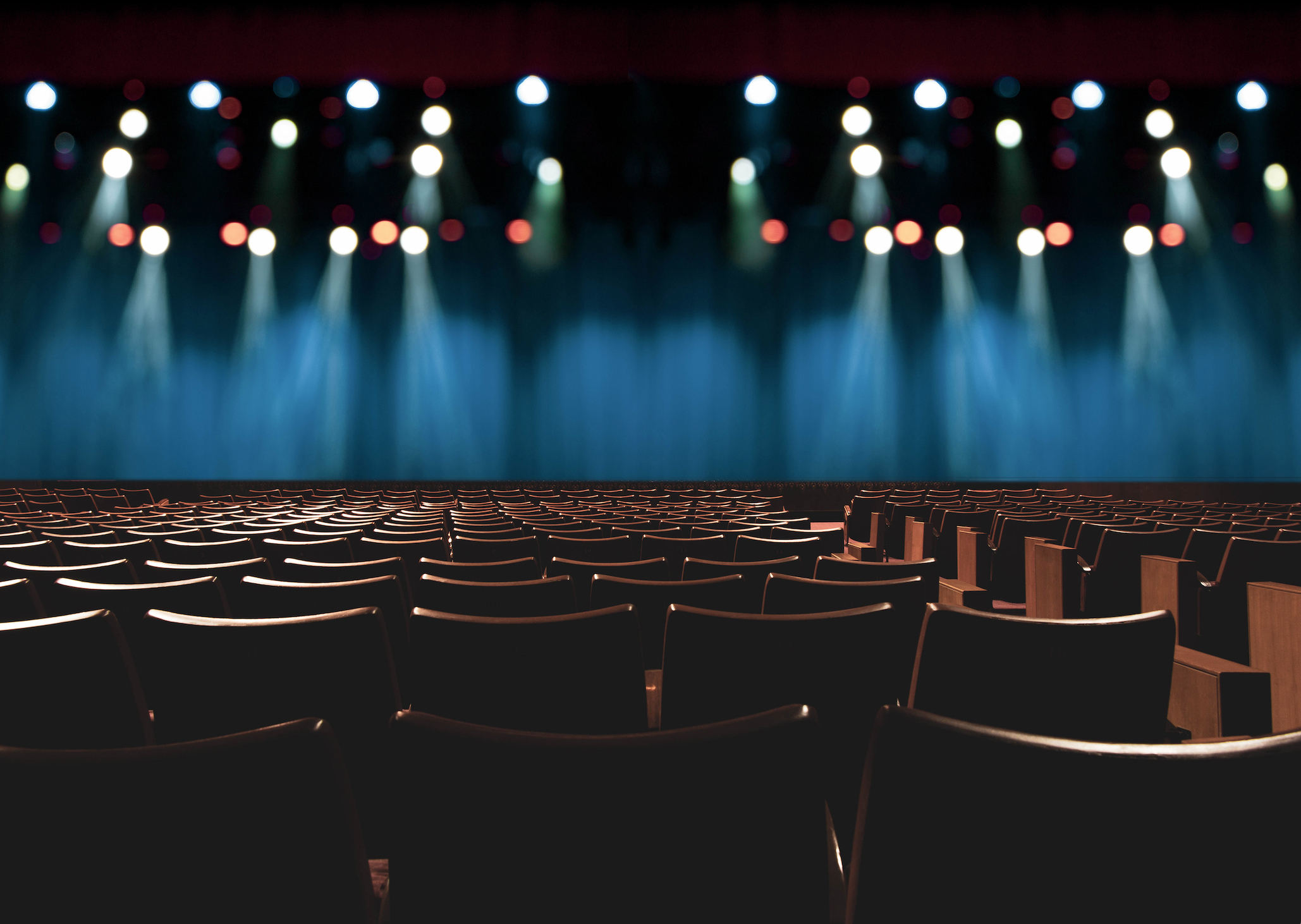 Empty rows of wooden theater seats looking onto a stage with blue lighting and white spotlights.