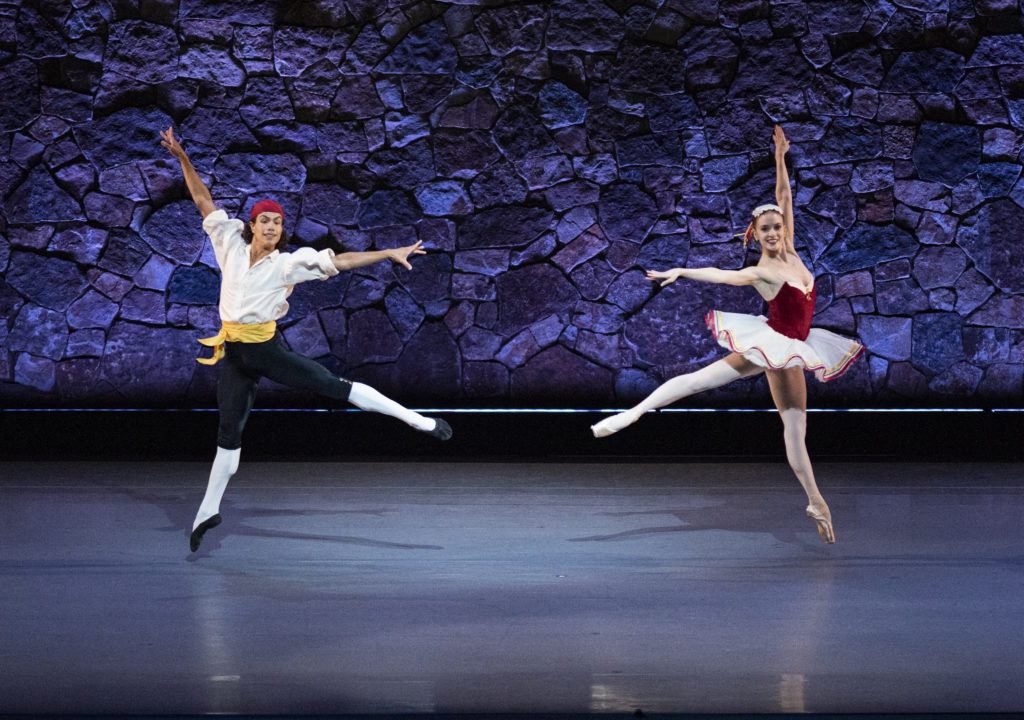 Esteban Hernández and Sasha de Sola perform first arabesque temps levés in opposite directions onstage, in front of a textured purple wall. Esteban wears a white peasant blouse, red bandana and black and white tights. Sasha wears a red and white tut, pink tights and pointe shoes and a white headpieces.