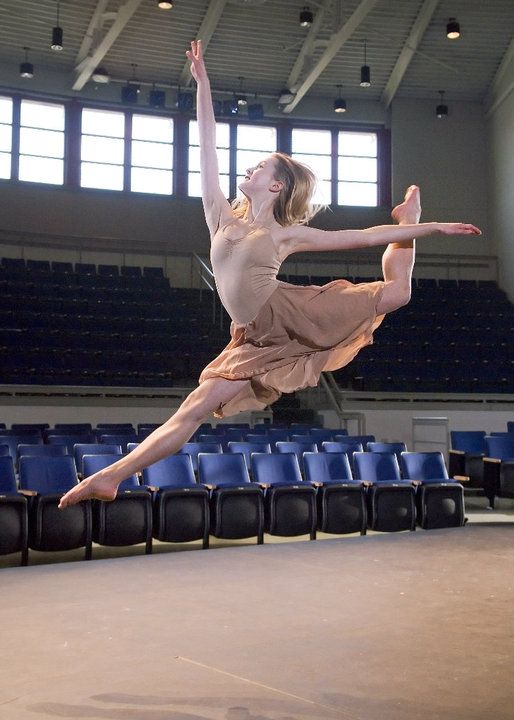 In a large auditorium, Grace Young performs a giant sissone fermé with her back leg bent in attitude and her arms in first arabesque. She wears a mauve dance dress and her long blonde hair down. Behind her are rows of auditorium seats.