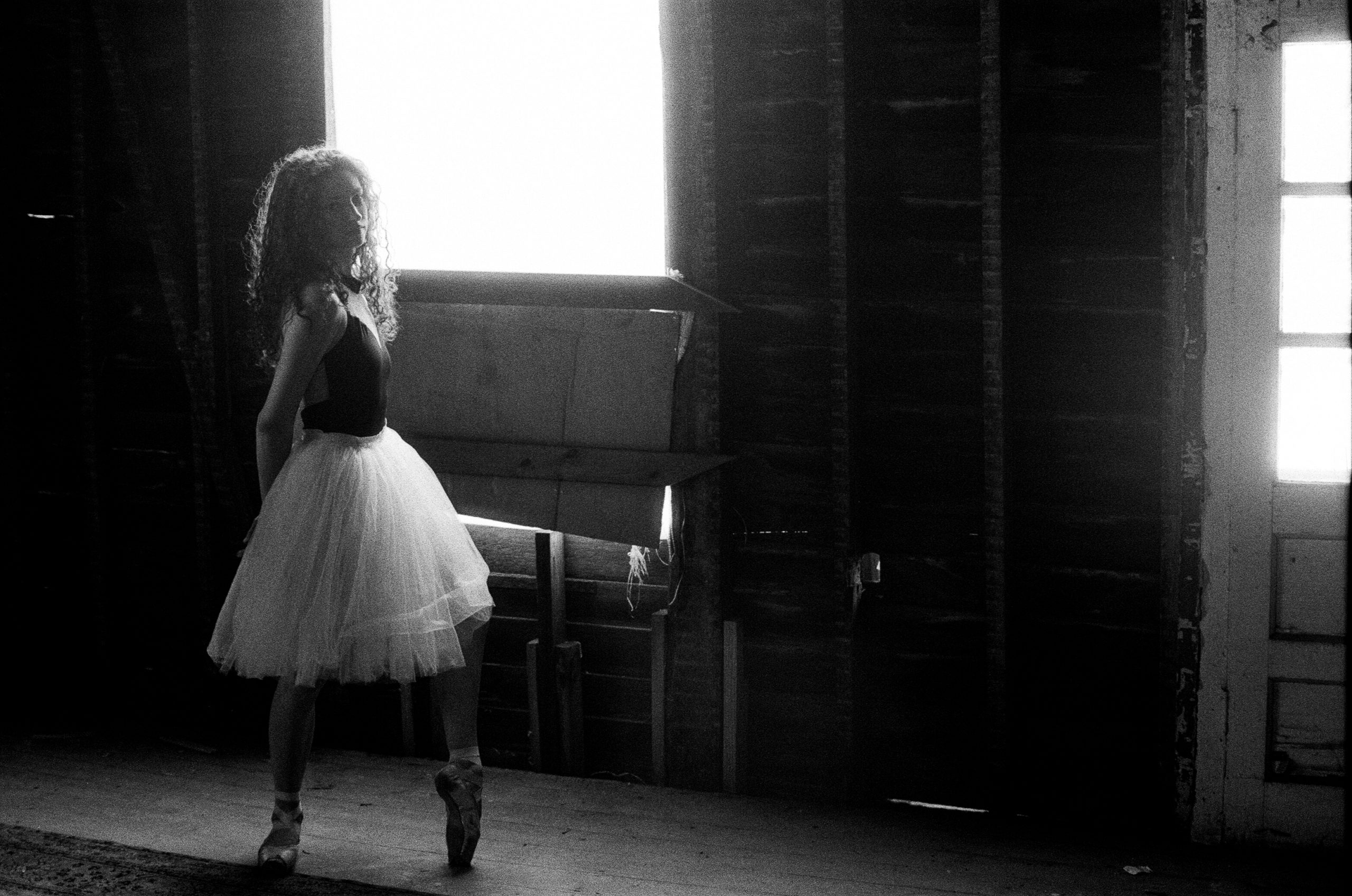 In this black and white photo, Anna Jensen wears a dark leotard, knee-length white tutu and pointe shoes and poses in what looks like the inside of a gutted house. She stands in profile with her left foot propped on pointe and her hands clasped behind her, looking wistfully towards the camera.