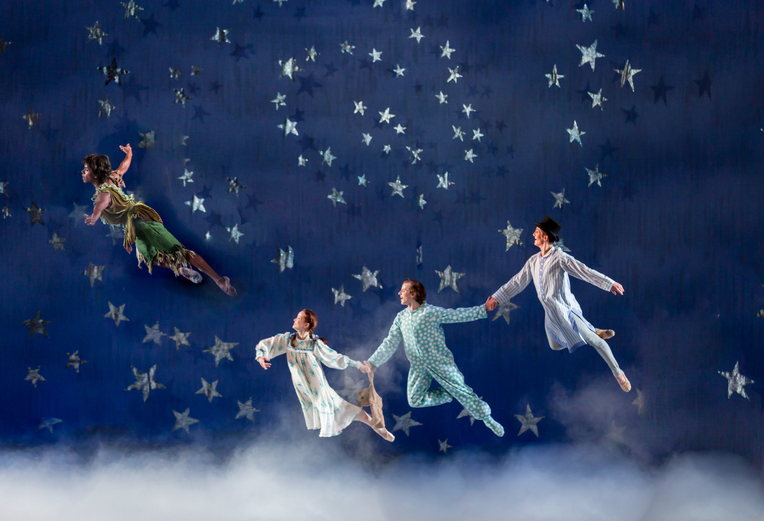 Three dancers dressed in costume as Peter Pan’s Wendy, Michael and John hold hands as they fly across a stage set with stars. Peter Pan also flies ahead of them.