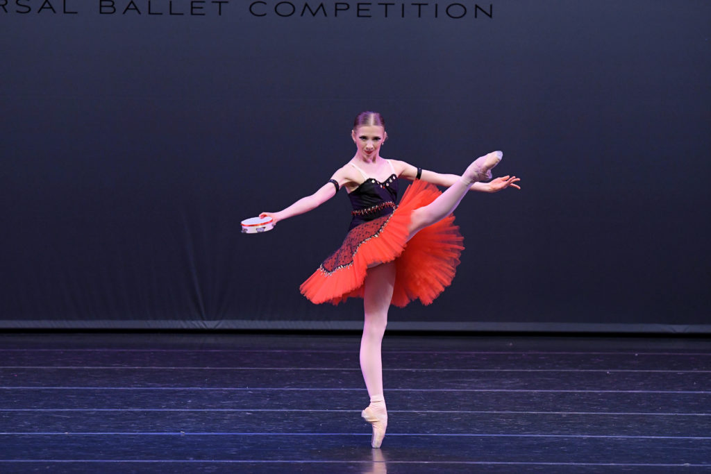 Julia Joyner wears a black and red tutu and holds a tambourine in her right hand. She stands on her left leg on pointe onstage and performs a battement at 90 degrees with her right leg, her arms in second, palms facing up.