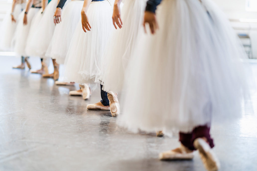 A photo shows a line of female dance dancers from the waist down, each wearing long Romantic-style practice tutus. They stand in b-plus with their left hands raised out of frame and their right hands in demi seconde.