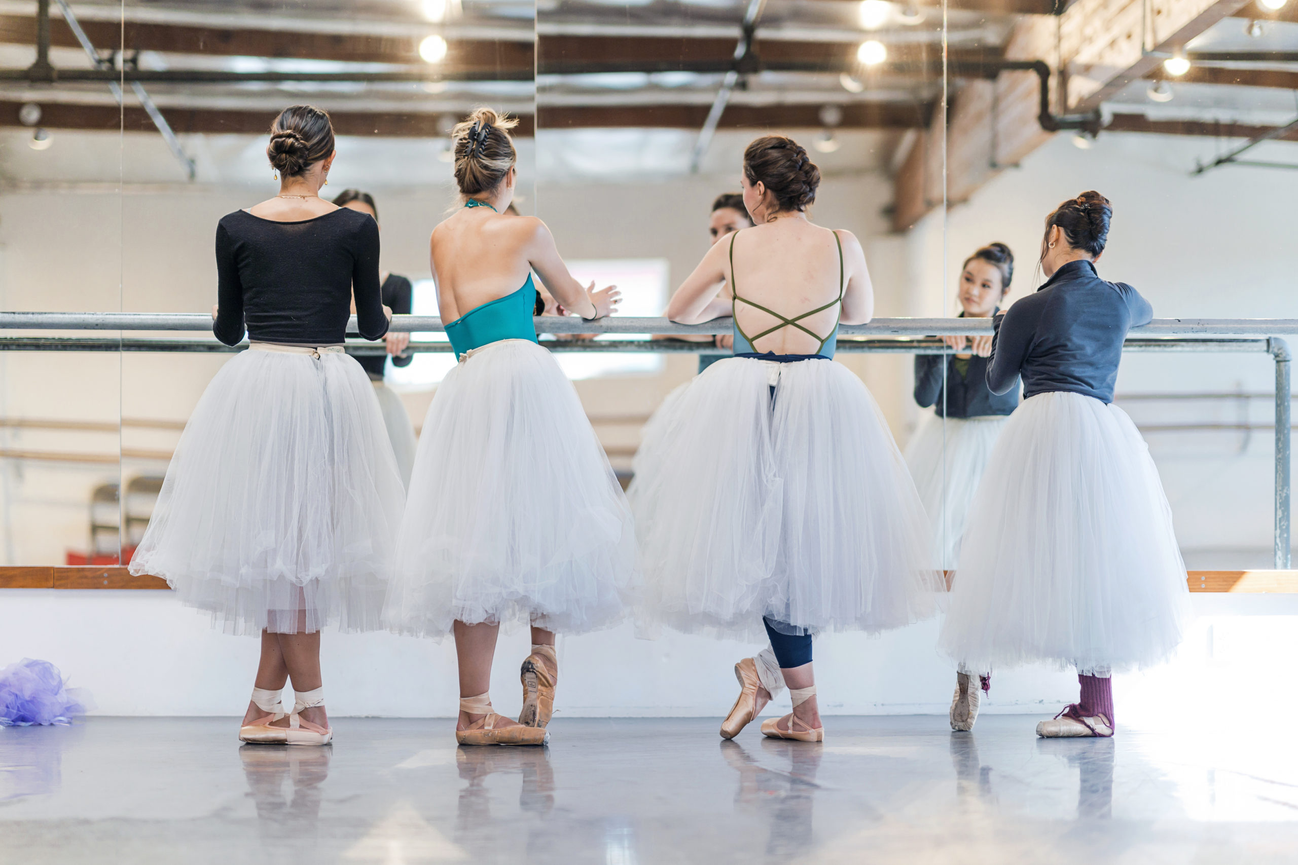 Four female dancers stand in a resting position facing the barre of a dance studio. The wall is a floor-to-ceiling mirror. They each wear different style leotards but all have matching white Romantic-length practice tutus.