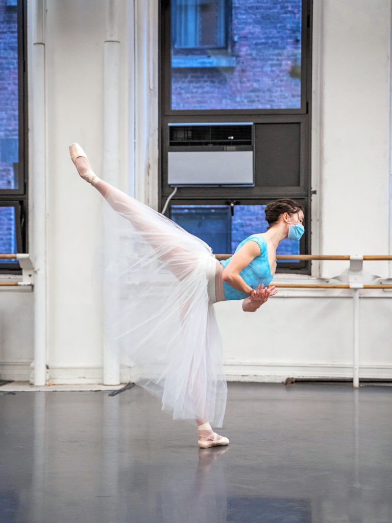 Cassandra Trenary practices a penché on her left leg, crossing her hands at her chest, during a Giselle rehearsal. She wears a blue leotard, blue face mask, pink tights and pointe shoes and a white romantic tutu.