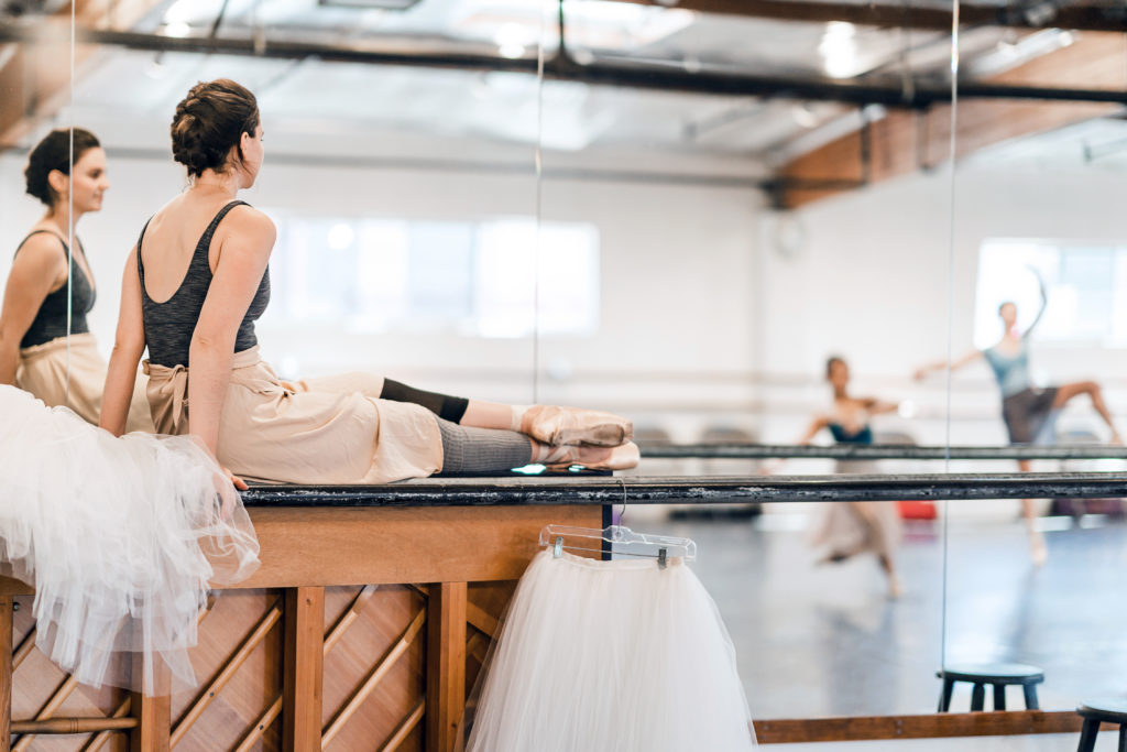 Shelby Whallon sits on top of a piano in the corner of a dance studio. She wears a dark leotard and a knee length skirt, footless tights and statin pointe shoes. She faces into a mirror, where you can see other dancers practicing in the studio behind her.
