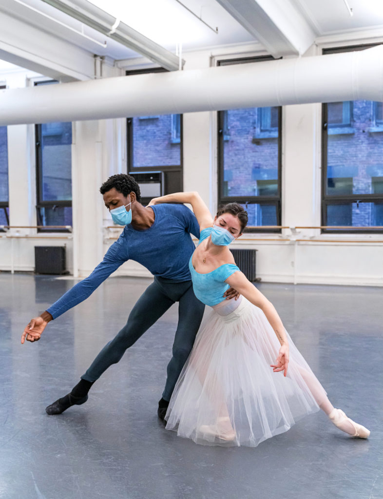 Calvin Royal III and Cassandra Trenary rehearse Giselle in a ballet studio. They stands next to each other hip to hip, with her right arm around his left shoulder and his left arm around her waist. They both bend their inside knee, with their outside leg in tendu, and open their free arms out. Trenary wears a blue leotard, white romantic tutu and pointe shoes, while Royal III wears a long-sleeved blue T-shirt, black tights, and black socks and ballet slippers. Both wear face masks.