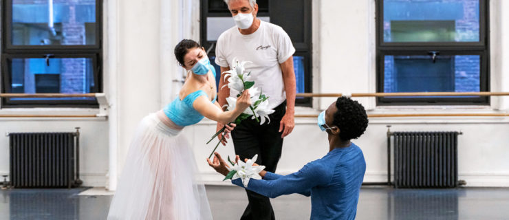 During a rehearsal at a dance studio, Kevin McKenzie, wearing a white T-shirt and black pants, stands behind Cassandra Trenary and Calvin Royal III and observes as they perform a scene from Giselle. Trenary, wearing a blue leotard, white romantic tutu and pointe shoes, stands with her feet loosely crossed and bends forward towards Royal III, who is facing her on both knees. He wears a blue T-shirt and black tights, and is catching the lily flowers that Trenary is dropping in his arms..