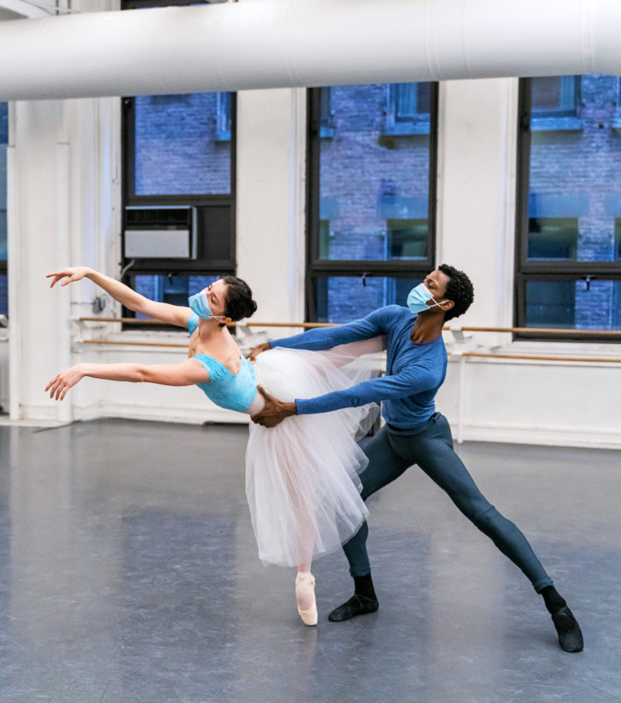 Calvin Royal III holds Cassandra Trenary by the waist as she does a fourth arabesque elongé on pointe, reaching her arms forward in front of her. They practice in a large dance studio with three large windows. Trenary wears a blue leotard, pink tights and pointe shoes and a white romantic tutu, while Royal III wears a long-sleeved blue T-shirt, black tights and black socks and ballet slippers. Both also wear face masks.
