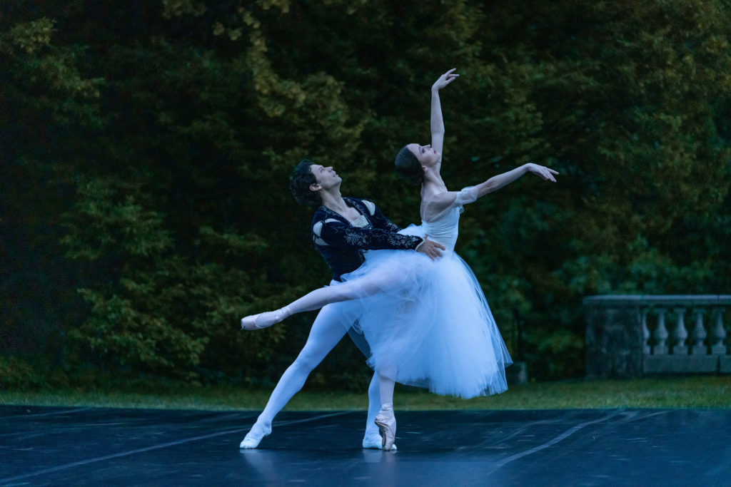 Herman Cornejo, in white tights and a dark velvet tunic, holds Skylar Brandt's waist as she stretches into a low third arabesque on pointe. She wears a long white Romantic tutu, and both dancers arch back slightly and look up towards the sky.