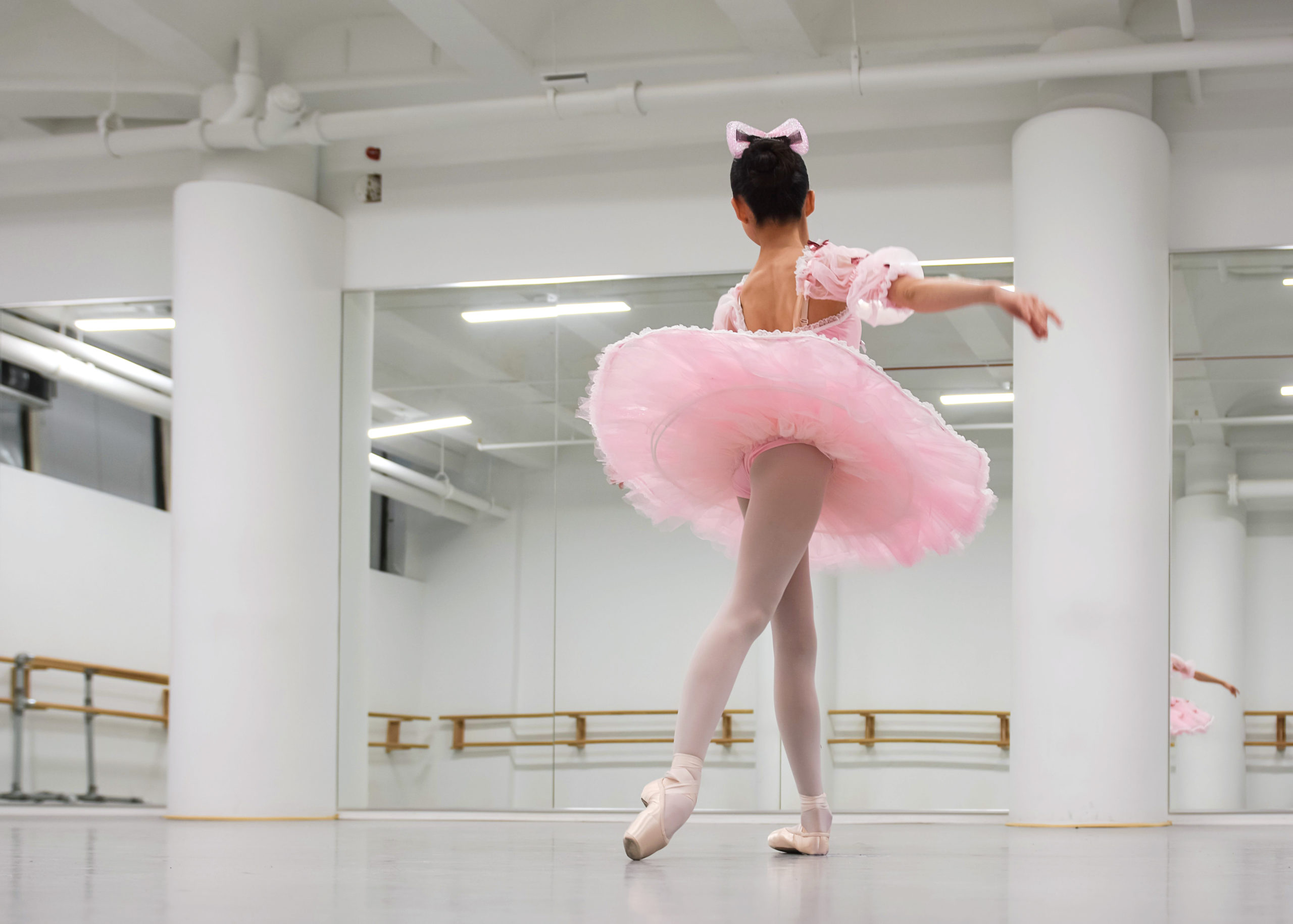 A young ballet student is shown from behind in a large dance studio. She wears a pink tutu, tights and pointe shoes and a bow in her hair, and stands in tendu croisé in derriere.