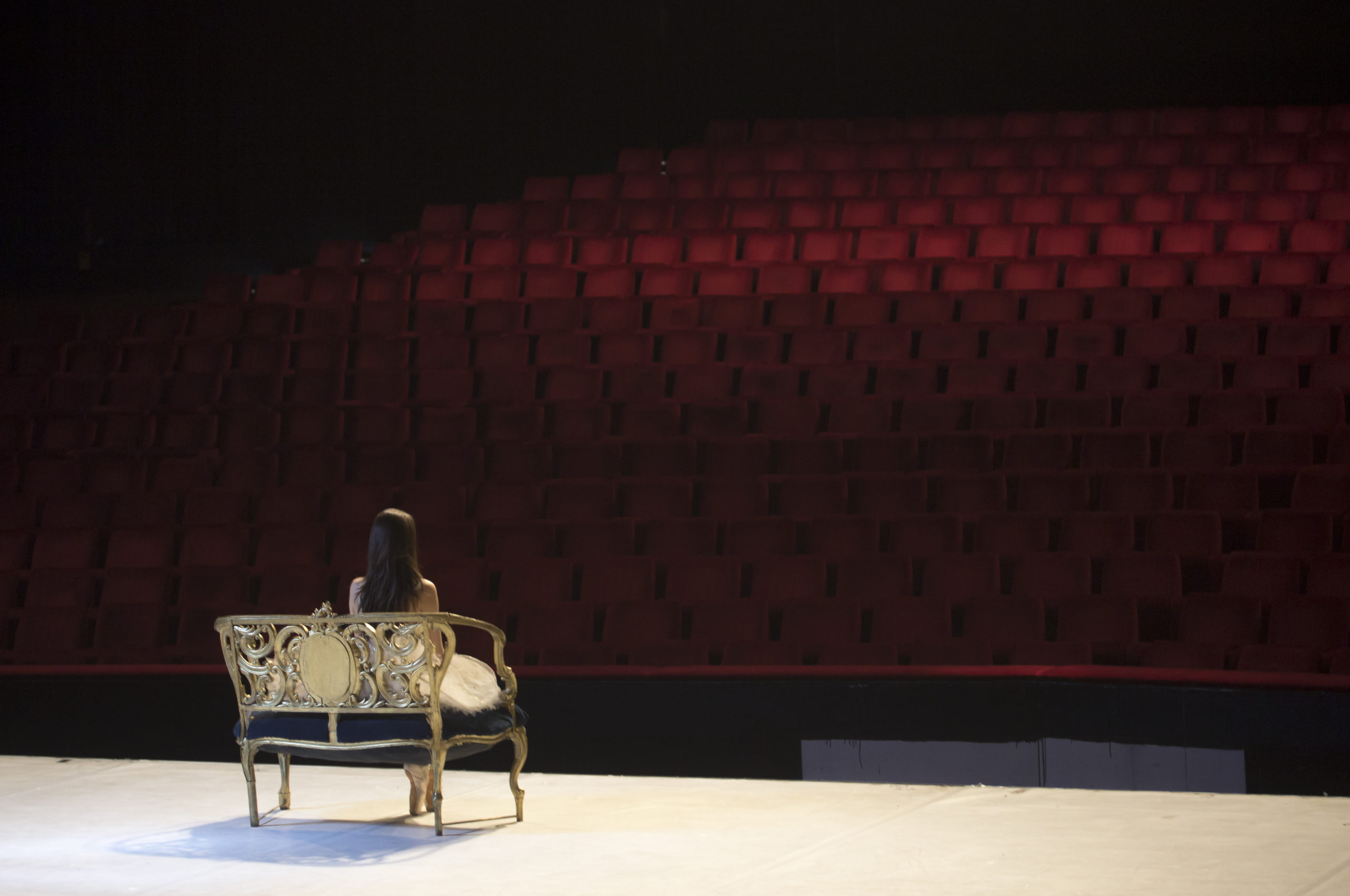 Young ballerina in a large theater is shown sitting on a bench in costume, looking out at the empty seats in front of her.