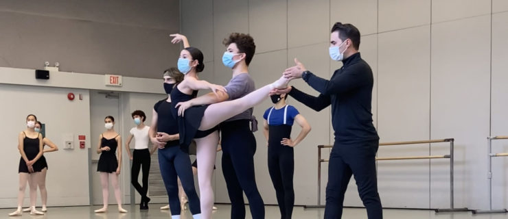 In a large dance studio, two young ballet students practice a pas de deux, with the girl on pointe in first arabesque and the boy standing behind her holding her waist. Joan Boada, wearing a black turtleneck, pants and tennis shoes and face mask, stands to the side and adjust the ballerina's lifted left foot. A group of ballet students stand to the right of them, watching.