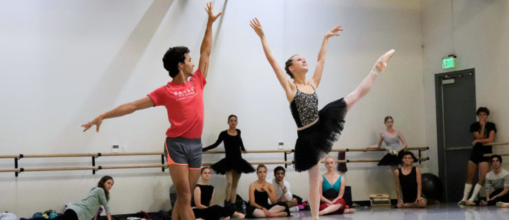 In a crowded dance studio, a male dancer stands in 5th position and releases Abby Phillips Maginity's hand as she executes a développé in á la second on pointe. The dancers all wear various dance clothes, and Abby wears a practice tutu.